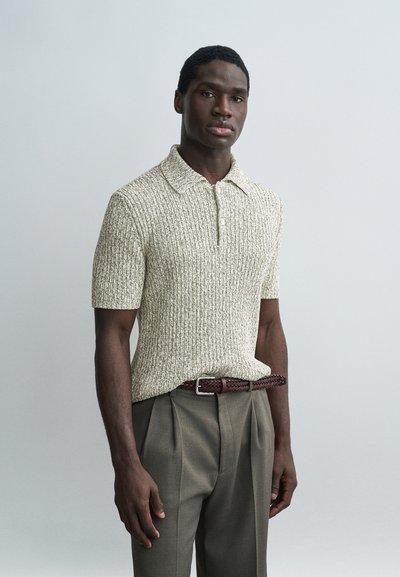 Homme portant un polo en maille beige texturé à manches courtes et un pantalon plissé olive avec une ceinture tressée marron, sur un fond uni.