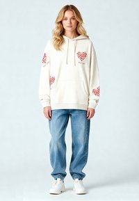 Krämfärgad hoodie med röda hjärtan på ärmarna, framfickan och text; matchad med ljusblå lösa jeans och vita sneakers.
