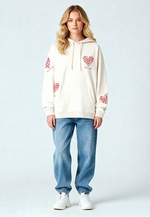 Crème hoodie met rode hartpatronen op de mouwen, een voorkantzak en tekst; gecombineerd met lichtblauwe losse spijkerbroeken en witte sneakers.