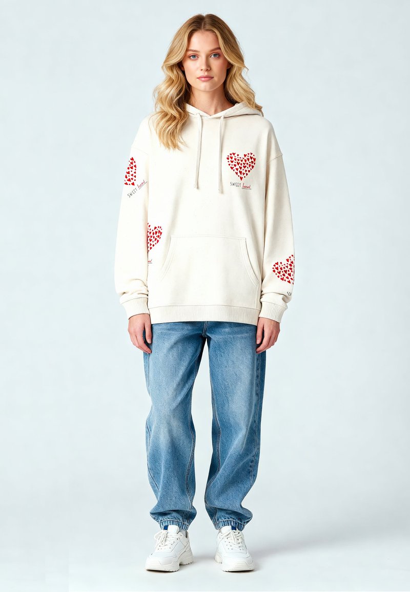 Krämfärgad hoodie med röda hjärtan på ärmarna, framfickan och text; matchad med ljusblå lösa jeans och vita sneakers.