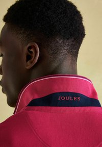 Muž s krátkými kudrnatými vlasy, oblečený v červeném polo tričku s vyhrnutým límcem a nápisem "JOULES" vyšitou na zadním límci.