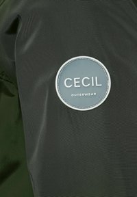 Donkergroene bovenkleding met een ronde grijze leren patch waarop "CECIL" en "OUTERWEAR" in het wit zijn geperst. Gladde, gestructureerde stof.