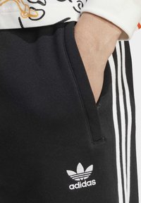 Handen i fickan på svarta Adidas träningsbyxor med vita ränder och logotyp, tillsammans med en vit ärm och ett mönstrat linne.