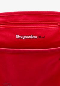 Raudonas audinio maišelis su užtrauktuku, ant priekio esantis baltas tekstinis logotipas "Visi yra Chuck" ir apvalus prekės ženklas.