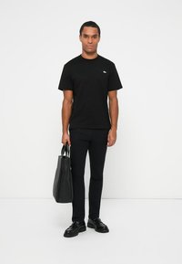 T-shirt noir à manches courtes avec un petit logo vert, associé à un pantalon slim noir et des chaussures noires, tenant un tote bag noir.