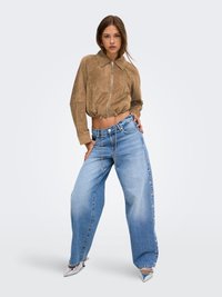 Femme portant une veste marron courte et un jean bleu à jambes larges avec des détails de boutons sur les côtés, posant avec une main sur la hanche.