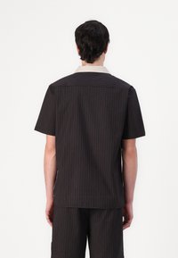 Camicetta a maniche corte in tessuto scuro con righe verticali. Presenta un colletto beige e una linea dritta, progettata con una silhouette semplice e pulita.
