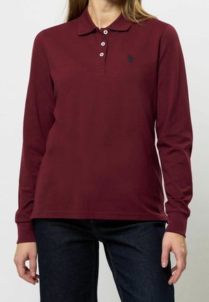 Woman wearing a long-sleeve maroon polo shirt with three buttons and dark jeans against a plain background.