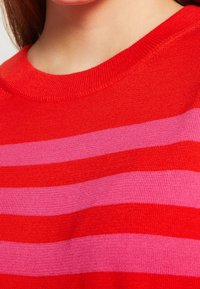 Punainen ja vaaleanpunainen raidallinen neulepaita, jossa on pyöreä pääntie, ribattu pinta ja vuorotellen leveät raidat, korostaen kirkaita värejä.