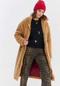 Žena v červené čepici, béžovém teddy kabátu, černém svetru s houbovými nášivkami a kalhotách s leopardím potiskem, pózuje na bílém pozadí.