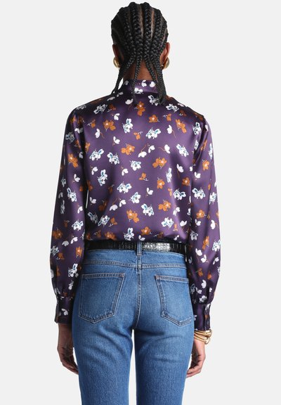 Personne aux cheveux tressés porte un chemisier violet à fleurs rentré dans un jean bleu taille haute avec une ceinture noire et des bracelets dorés au poignet.