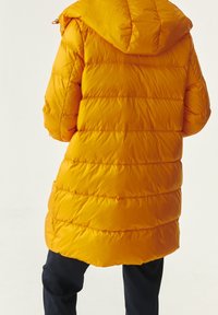 Chaqueta de abrigo amarilla con capucha, que presenta un diseño acolchado y un corte ligeramente más largo. Textura suave con costuras y costurados visibles.