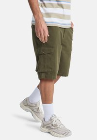 Man in olijfgroene cargoshorts, witte crew sokken, beige en witte sneakers, staand met één hand in zijn zak en de andere aan de zijkant.