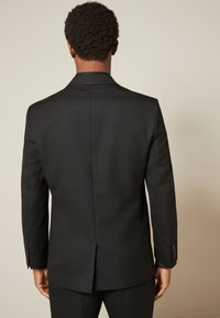 Veste de costume noire à texture lisse, avec une fente unique au dos et des poignets boutonnés. Caractérisée par une coupe ajustée et un design structuré.