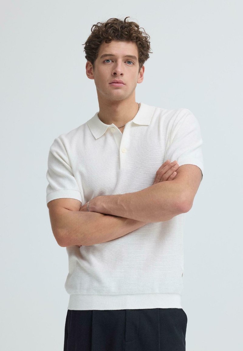 Jeune homme aux cheveux bouclés portant un polo blanc à manches courtes et un pantalon foncé, debout les bras croisés devant un fond uni.