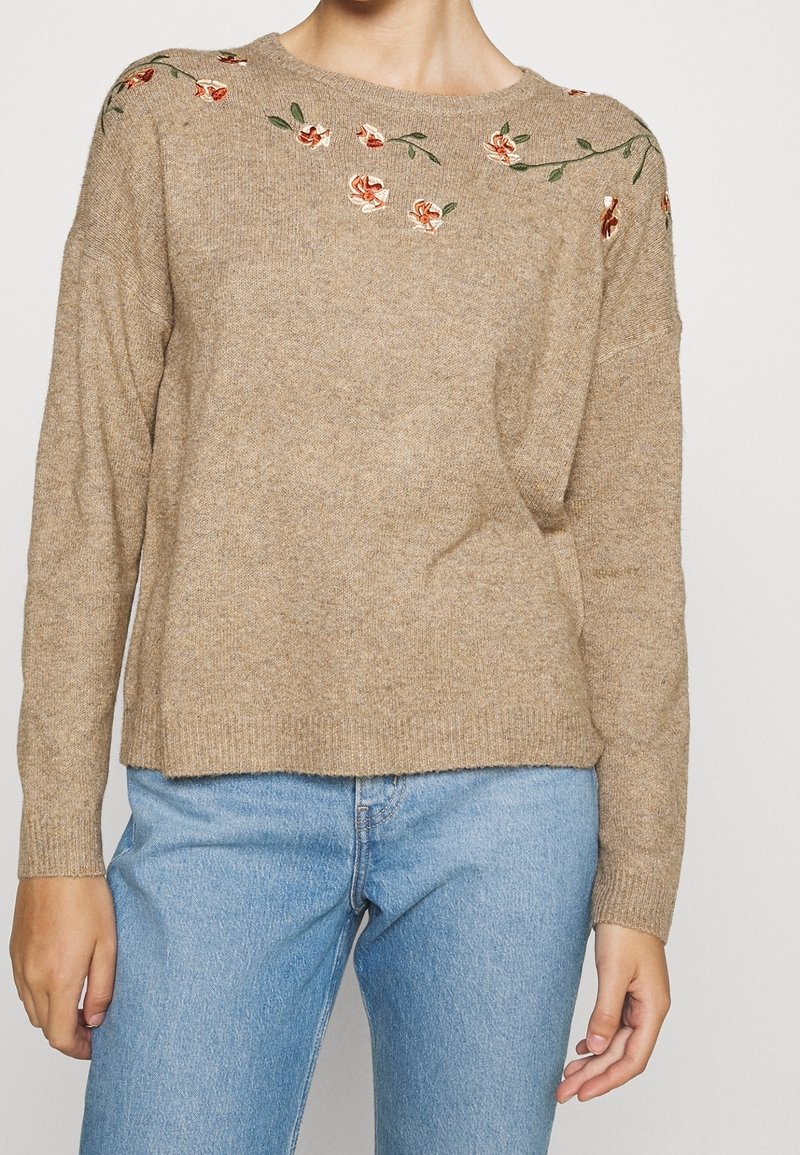 Femme portant un pull en maille beige avec de petites broderies florales rouges sur les épaules, associé à un jean denim bleu clair.