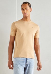 Camiseta de manga corta y cuello redondo en algodón beige. Presenta un sutil logo en el pecho, combinada con jeans de mezclilla azul claro.