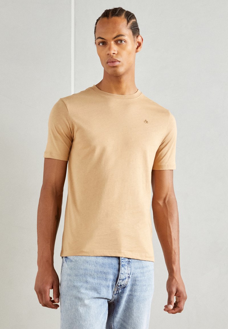 Camiseta de manga corta y cuello redondo en algodón beige. Presenta un sutil logo en el pecho, combinada con jeans de mezclilla azul claro.