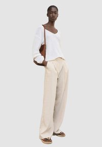 Haut léger à manches longues blanc, pantalon large plissé beige, et sac à bandoulière tissé marron. Dessandales à bout ouvert complètent la tenue.