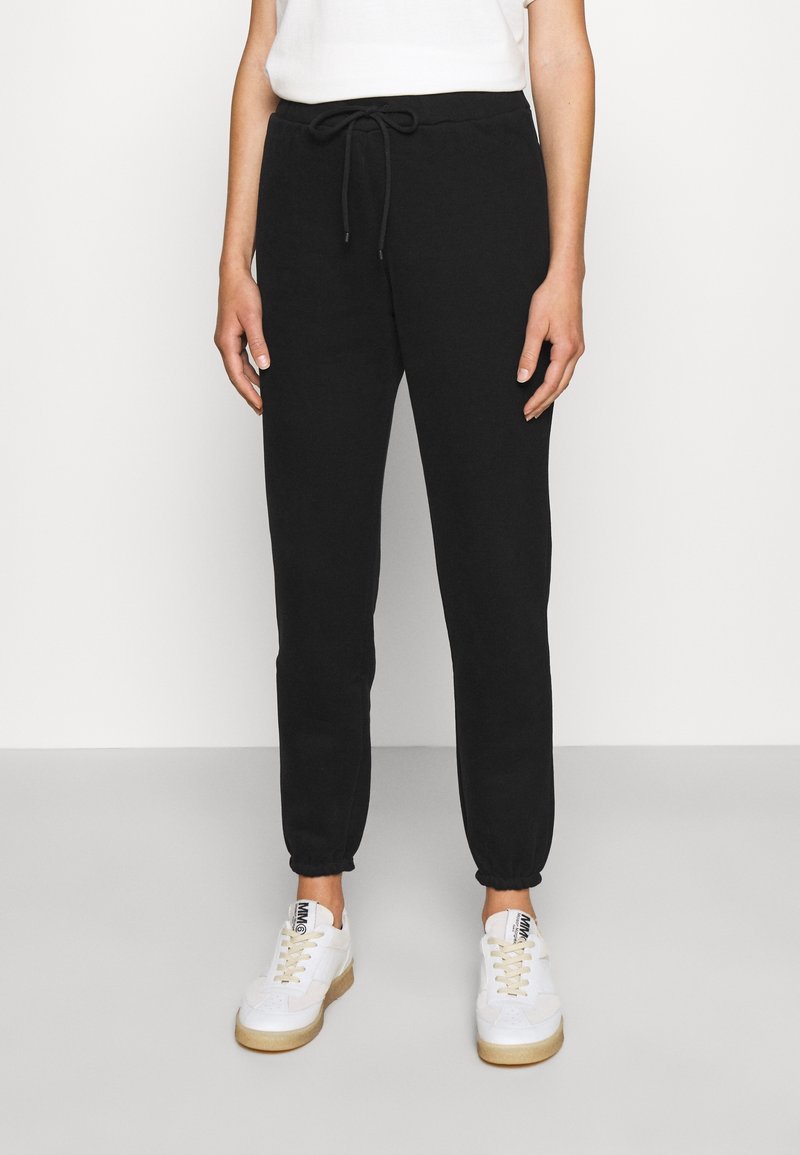 Person med svarta joggers med dragsko och vita sneakers, stående mot en ljus, enfärgad bakgrund.