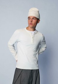 Weißes, geripptes Langarmshirt mit Henley-Ausschnitt und Knopfleiste, kombiniert mit einer strukturierten weißen Mütze und grauen Nadelstreifenhosen.