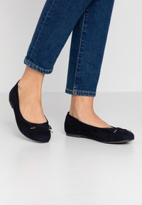 Füße in dunkelblauen Wildleder-Ballerinas mit kleinen Schleifenakzenten, kombiniert mit cropped blauen Jeans vor einem schlichten hellen Hintergrund.