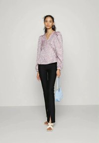 Mulher em blusa de manga longa floral, calças pretas justas com fendas frontais, sandálias brancas, segurando uma pequena bolsa azul-claro, em pé contra uma parede simples.