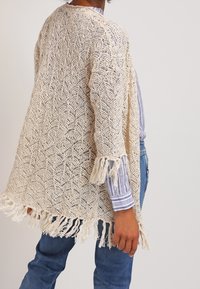 Open-knit cardigan in crème, met een scalloped patroon, franjes aan de randen en een losse snit. Gedirigeerd over een gestreept topje met blauwe jeans.