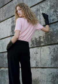 Joven mujer con cabello rizado rubio que lleva una camiseta corta rosa, pantalones negros y guantes negros, apoyada contra una pared de piedra.