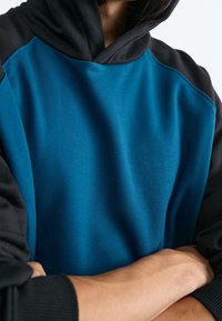 Persona che indossa un felpa blu e nera con le braccia incrociate, torace visibile e viso parzialmente fuori dal riquadro.