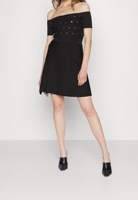 Robe noire décolletée avec un corsage ajusté mettant en valeur des accents métalliques ; jupe évasée. Portée avec des mules noires à bout ouvert.