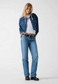 Spijkerjack en blauwe spijkerbroek met een aansluitend wit overhemd. Het jack heeft zakken met knopen en een capuchon, met een bruine riem en zwarte laarzen.