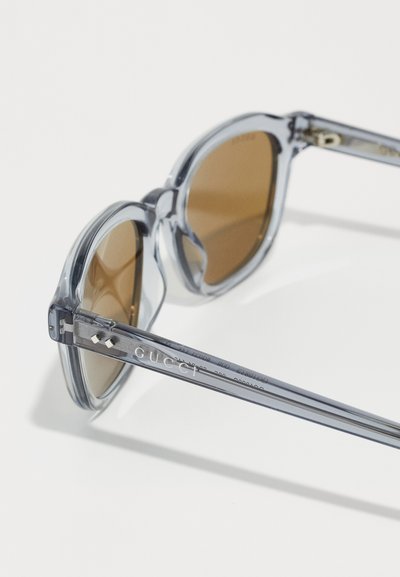 Klare Plastiksonnenbrille mit braunen Gläsern, runder Form und grauem Rahmen. Mit "Gucci"-Logo am Schenkel. Metallverzierungen sichtbar.