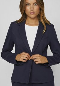 Mørkeblå blazer med figursyet pasform, enkeltknaplukning og to forlommer. Glat stof med let glans. Hvid top under.