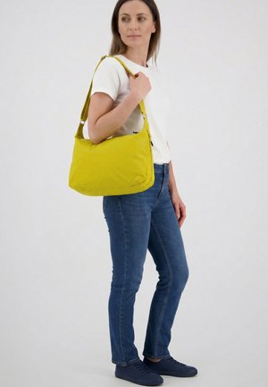 Woman in white shirt and jeans carries yellow shoulder bag, standing against plain light background, looking to side.