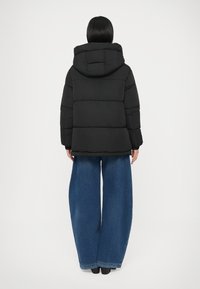 Must kapuutsiga puffer-jakk, millel on tikandiga disain ja kasti kujuga lõige, kombineeritud laia säärega siniste teksapükstega.