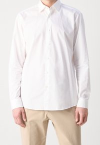 Camicia bianca a maniche lunghe in cotone. Presenta un colletto classico, chiusura con bottoni e una texture liscia. Indossata con pantaloni beige.