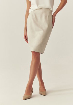 Kobieta ubrana w beżową ołówkową spódnicę do kolan, białą włożoną do środka bluzkę oraz beżowe szpilki z zaokrąglonym noskiem, stoi ze skrzyżowanymi nogami.
