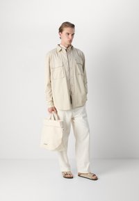 Lichtbeige overhemd met knopen en twee borstzakken, gecombineerd met lichtgekleurde losse broeken en platte sandalen. Draagt een lichtbeige schoudertas.
