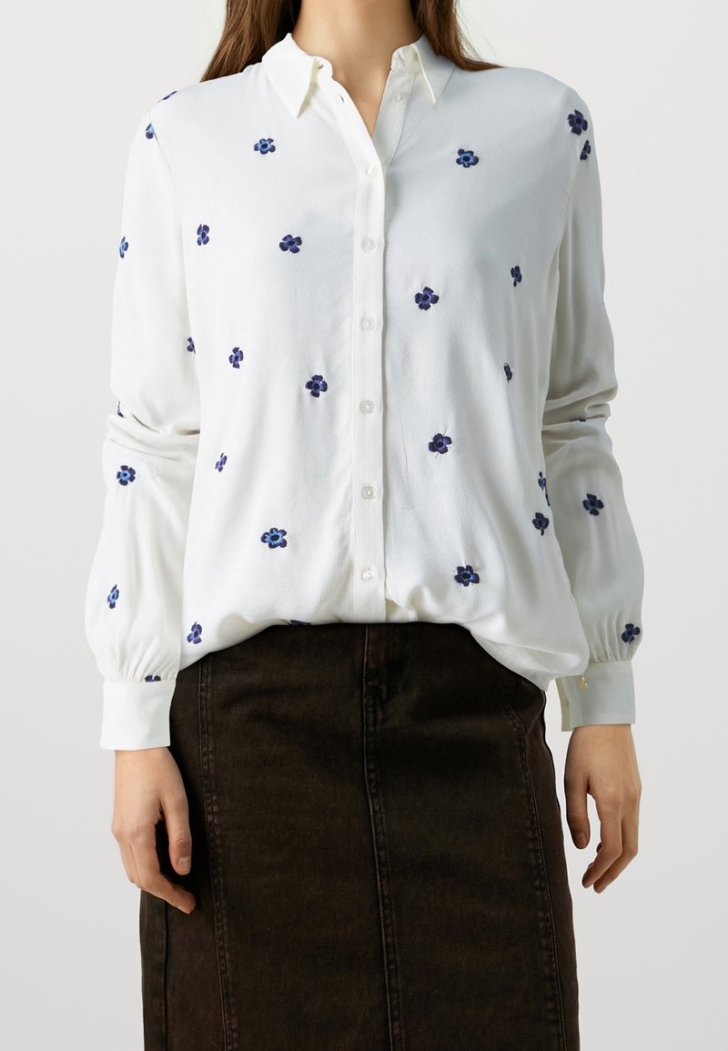Femme portant un chemisier blanc boutonné avec des imprimés floraux bleus et une jupe sombre, debout devant un fond clair uni.