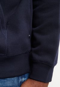 Felpa blu navy con tessuto testurizzato, polsini a coste e dettagli di cucitura laterale. Include una piccola etichetta del marchio sull'orlo.