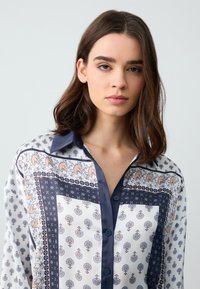 Chemisier blanc avec des accents marins à motifs. Présente des motifs paisleys et floraux, un col et une fermeture à boutons. Tissu lisse et léger.
