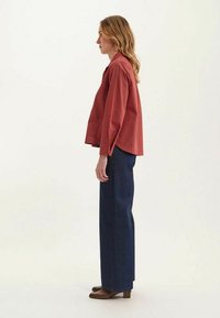 Chemise à carreaux rouges à manches longues, associée à un pantalon en denim foncé taille haute et des bottines à talons marron, vue de côté.
