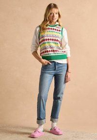 Grüner Strickwesten mit bunten Mustern über einem weißen Hemd, kombiniert mit blauen Jeans und rosa Holzclogs mit grünen Akzenten. Glatte Textur.