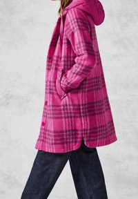 Abrigo a cuadros rosa con capucha, con líneas grises contrastantes, dos bolsillos laterales y una tela de textura suave. La longitud llega a media pantorrilla.