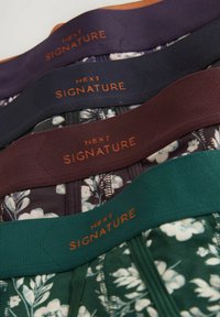 Quatre sous-vêtements à motif floral avec des ceintures élastiques larges en violet, marine, bordeaux et vert, chacun étiqueté « Next Signature » en orange.