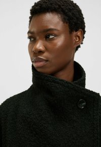 Czarny, teksturowany płaszcz z wysokim kołnierzem, z dużym czarnym zapięciem na guzik. Materiał wydaje się miękki i gruby, odpowiedni na zimną pogodę.