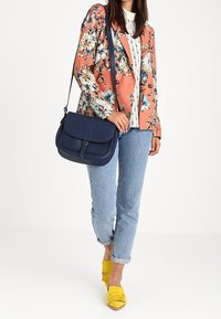 Borsa a tracolla blu navy con patta, texture liscia e dettagli di cucitura. Indossata con un blazer floreale pesco, camicia a righe e scarpe gialle con frange.