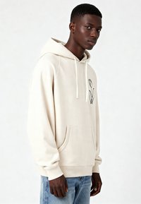 Sweat à capuche beige en tissu doux, avec une poche kangourou, une capuche à cordon de serrage et un motif de contour de fleur noire sur le côté gauche de la poitrine.