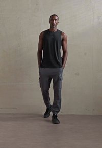 Hombre atlético caminando en interiores, vistiendo una camiseta sin mangas negra, pantalones joggers gris de carga y zapatillas negras, con un fondo de pared de concreto liso.